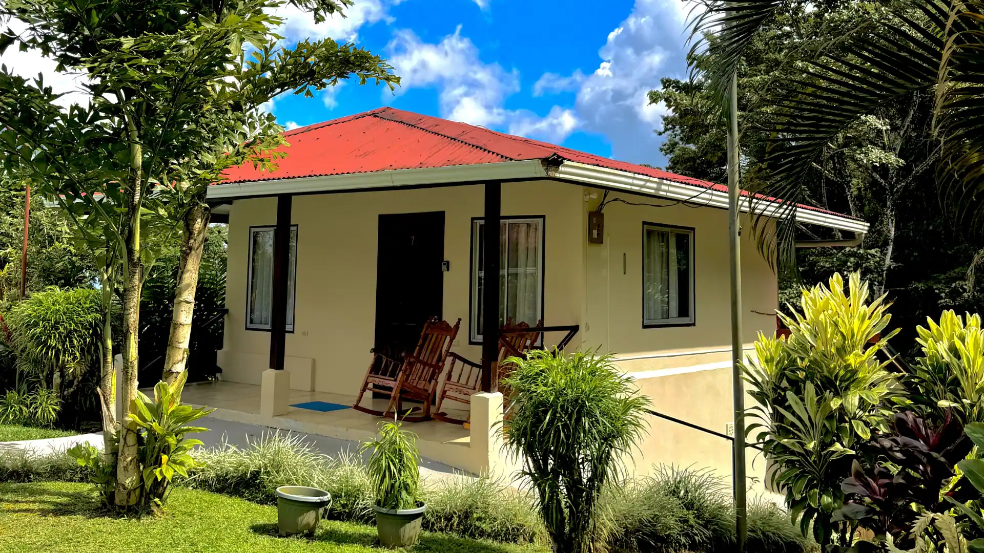Cabaña de color crema con techo rojo, terraza frontal con mecedoras de madera y jardines tropicales bajo un cielo azul con nubes.