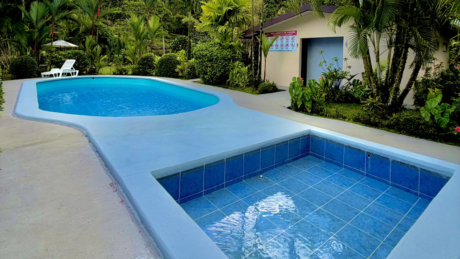 Piscina curva y jacuzzi cuadrado integrados en un jardín tropical con reglamento de uso visible al fondo.
